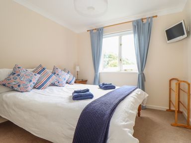 A bedroom with a bed with patterned pillows folded towels a bedside table with lamp blue curtains a window and a wall-mounted television at Deja Blue in Falmouth