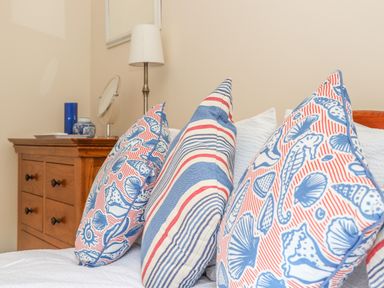 Three decorative pillows with seashell and striped patterns on a bed next to a wooden dresser with a lamp and mirror at Deja Blue in Falmouth