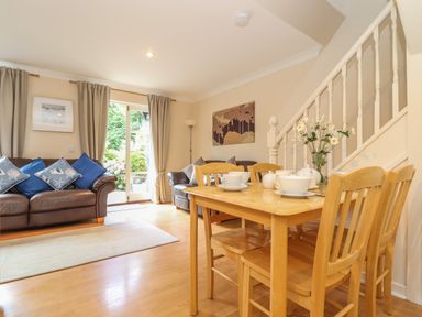 A dining area with a wooden table and chairs set with tea cups next to a staircase and a living room with two sofas and a door leading outside at Deja Blue in Falmouth