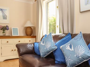 A living room with a brown leather sofa and blue cushions next to a window and a white sideboard with a lamp and flowers at Deja Blue in Falmouth