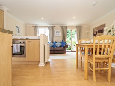 An open-plan room with a kitchen area, wooden dining table with chairs, and a seating area with a sofa and cushions at Deja Blue in Falmouth