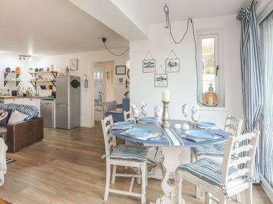 A dining area with a table set for six with blue plates and striped cushions next to a kitchen with shelves and appliances at The Old Boat Store in Morfa Nefyn