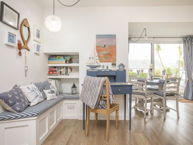 A corner seating area with cushions and bookshelves next to a blue desk and wooden chair with a blanket by a dining table with chairs near large windows at The Old Boat Store in Morfa Nefyn
