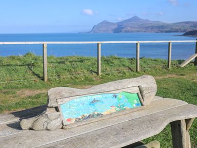 A wooden bench with an informational plaque and a coastal landscape with a fence and mountains in the background at The Old Boat Store in Morfa Nefyn