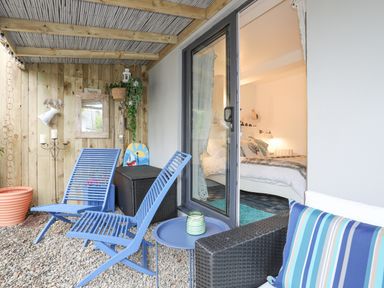 A covered outdoor seating area with blue chairs a small table and a sliding glass door leading to a bedroom at The Old Boat Store in Morfa Nefyn