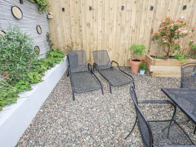 An outdoor patio area with two black metal lounge chairs a black metal table and chairs gravel flooring plants in a raised wooden planter and a wooden fence at The Old Boat Store in Morfa Nefyn