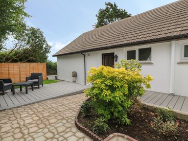 An outdoor patio area with a wooden deck and black patio furniture next to a white house with a brown tiled roof and garden bed at Little Valley View in Angarrack