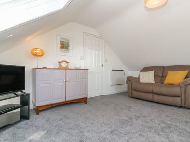 A carpeted room with a brown sofa with two cushions a white door a cabinet with a lamp clock and glass dish and a TV on a black stand at Little Valley View in Angarrack