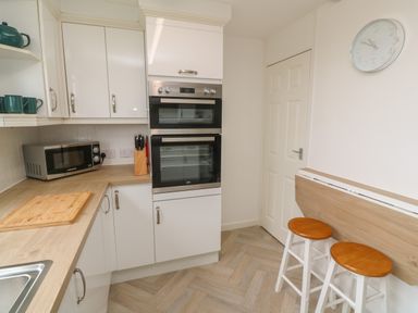 A kitchen with white cabinets wooden countertop double oven microwave knife block wall clock and two wooden stools at Little Valley View in Angarrack