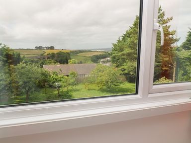 A view through a window showing trees a house with a tiled roof and distant fields at Little Valley View in Angarrack