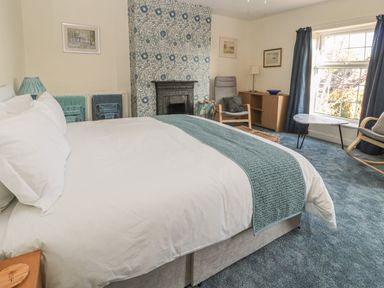 A bedroom with a large bed blue throw and cushions a chair a small table a fireplace and a window with curtains at Wordsworth Cottage West in Longframlington