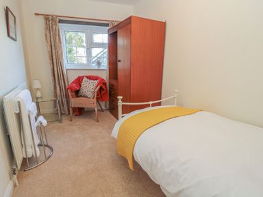 A bedroom with a single bed with a yellow throw a wooden wardrobe a wicker chair with a red blanket and a small table with a lamp at Wordsworth Cottage West in Longframlington