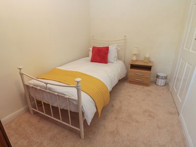 A bedroom with a single bed with white bedding and a yellow throw and red pillow next to a wooden nightstand with a lamp and tissue box at Wordsworth Cottage West in Longframlington