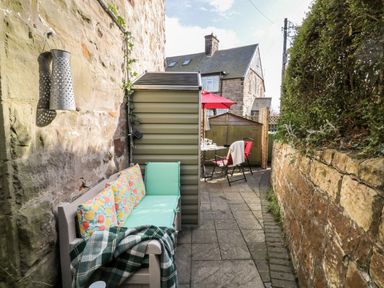 An outdoor seating area with a bench, cushions, a blanket, a table with chairs, an umbrella, and stone walls at Wordsworth Cottage West in Longframlington