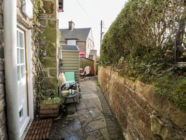 A narrow outdoor patio with a stone wall on one side seating and a small table near a door at Wordsworth Cottage West in Longframlington
