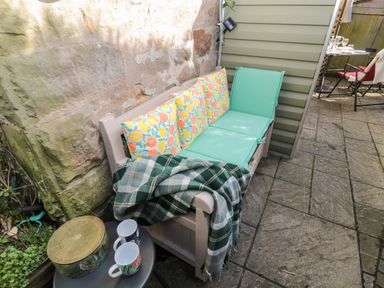 An outdoor seating area with a bench with cushions and a blanket next to a small table with two mugs and a tin at Wordsworth Cottage West in Longframlington