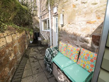 A narrow outdoor stone patio with a bench with cushions and a blanket at Wordsworth Cottage West in Longframlington