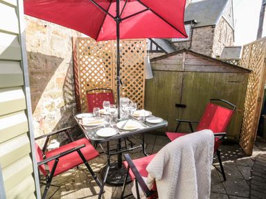An outdoor patio area with a table set for four under a red umbrella and red chairs at Wordsworth Cottage West in Longframlington