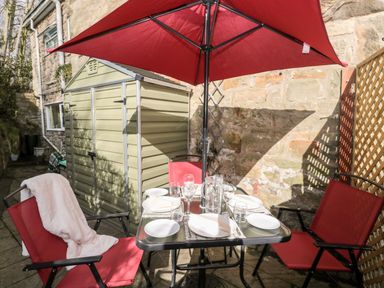 An outdoor patio area with a table set for four under a red umbrella next to a small shed at Wordsworth Cottage West in Longframlington