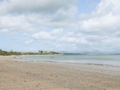 A sandy and pebbly beach with people near the shore and green hills in the background at Songbird Hideaway in Pwllheli