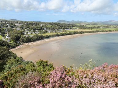 An aerial view of a curved sandy beach with people on the sand and in the water with houses and fields in the background at Songbird Hideaway in Pwllheli