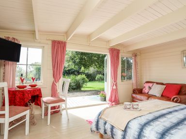 A room with a brown leather sofa a bed with cups on a tray a table with two chairs and red curtains at Songbird Hideaway in Pwllheli