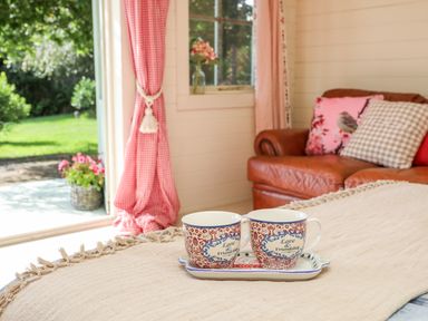 Two cups on a tray on a table with a blanket near a leather sofa and a window with red curtains at Songbird Hideaway in Pwllheli