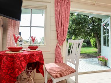 A dining area with a red tablecloth on a round table set with plates and wine glasses next to a white chair with a red checkered cushion at Songbird Hideaway Pwllheli
