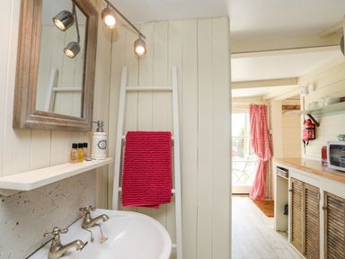 A bathroom sink with a mirror and a red towel on a white rack next to a kitchen area with wooden cabinets and red checkered curtains at Songbird Hideaway in Pwllheli