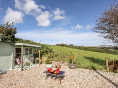 A gravel patio with a table and chairs beside a small shed and a green field in the background at Songbird Hideaway in Pwllheli