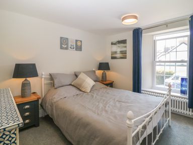 A bedroom with a bed a bedside table with lamps a window with curtains and wall art at Rosebank Cottage in Mortehoe