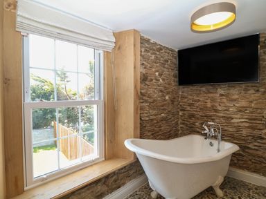 A bathroom with a clawfoot bathtub under a wall-mounted television and a window with a Roman shade at Rosebank Cottage in Mortehoe