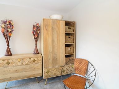 A room with wooden furniture including a chest of drawers and a cabinet with woven baskets and a chair with a metal frame and brown padded seat at Rosebank Cottage in Mortehoe