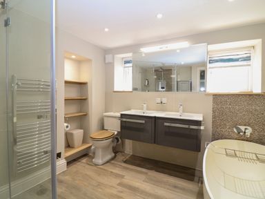 A bathroom with a shower, toilet, double sink, bathtub, and wooden shelves at Rosebank Cottage in Mortehoe