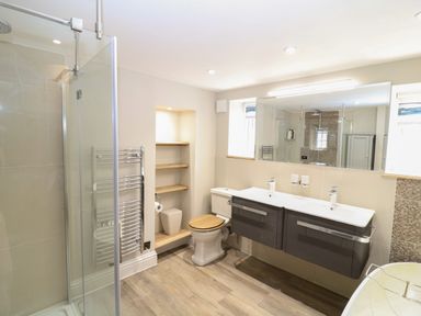 A bathroom with a shower enclosure toilet double sink vanity wooden shelves and a window at Rosebank Cottage in Mortehoe