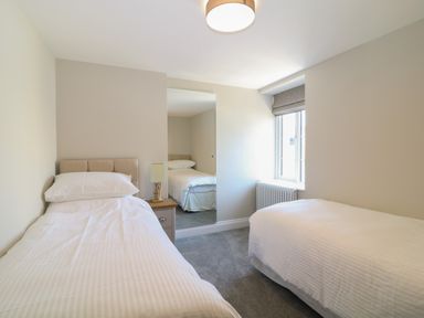A bedroom with two single beds, a bedside table with a lamp, a window with a blind, and a mirror on the wall at Rosebank Cottage in Mortehoe