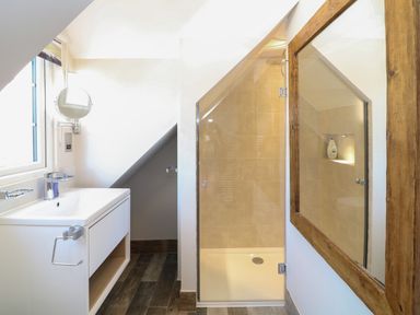 A bathroom with a white sink vanity under a window a glass-enclosed shower with beige tiles and a large wooden framed mirror at Rosebank Cottage in Mortehoe