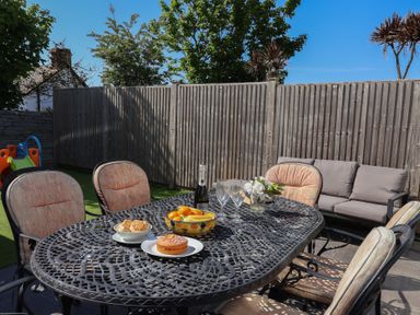 An outdoor patio with a metal dining table and cushioned chairs with fruit and cake on the table and a sofa against a wooden fence at Rosebank Cottage in Mortehoe