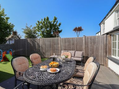 A patio with a metal table and six cushioned chairs next to a wooden fence with trees and play equipment in the background at Rosebank Cottage in Mortehoe