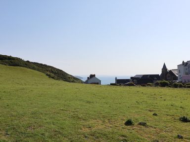 A grassy field with a hill on the left and houses with a church tower on the right near the coastline at Rosebank Cottage in Mortehoe