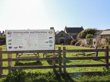 An information board about Woolacombe and Mortehoe behind a wooden fence with a museum building and garden in the background