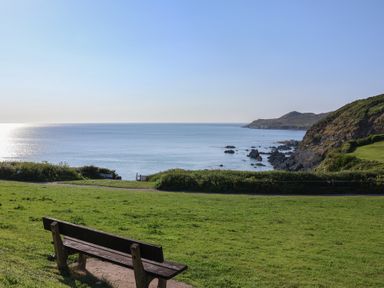 A bench on grass overlooking a calm sea and rocky coastline at Rosebank Cottage in Mortehoe
