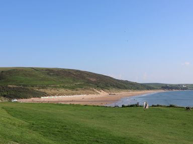 A grassy area overlooking a sandy beach with colorful beach huts and hills in the background at Rosebank Cottage in Mortehoe