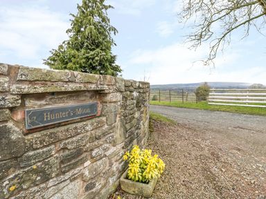 A gravel path next to a stone wall with a sign that says Hunters Moon and a planter with yellow flowers at Hunters Moon in Tenbury Wells