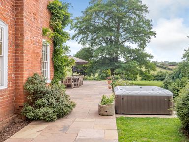 An outdoor patio with a hot tub wooden dining table chairs umbrella and large tree at Hilltop House in Tenbury Wells