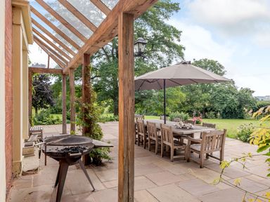 An outdoor patio with a wooden dining table and chairs under an umbrella and a barbecue grill near a glass-roofed pergola with trees in the background at Hilltop House in Tenbury Wells