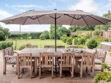 An outdoor patio with a wooden table and chairs under a large beige umbrella in a garden at Hilltop House in Tenbury Wells