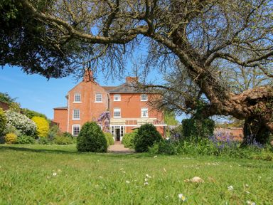 A large brick house with multiple windows and a garden with bushes flowers and a large tree at Hilltop House in Tenbury Wells