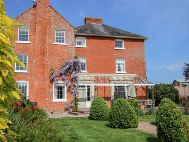 A red brick house with white framed windows and a glass patio cover surrounded by a garden at Hilltop House in Tenbury Wells