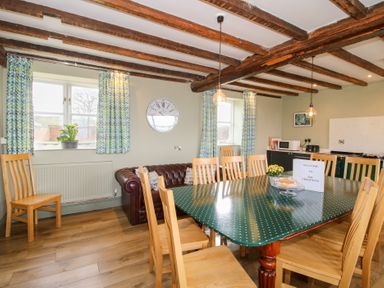 A dining room with wooden chairs around a large table and a leather sofa under windows with patterned curtains at The Chestnuts in Tenbury Wells